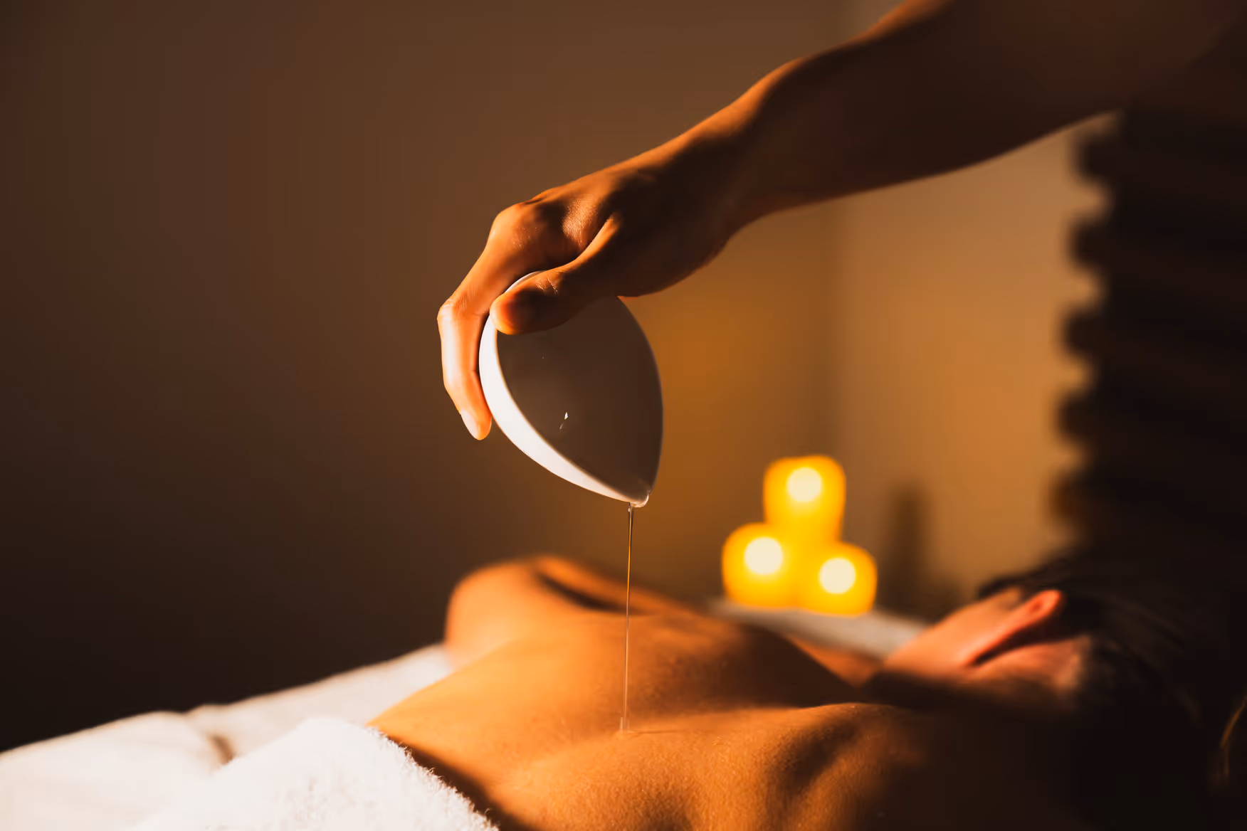 Main versant de l'huile chaude sur le dos nu d'une personne allongée lors d'un massage myBlend avec bougies allumées en arrière-plan.